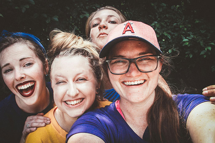Four women smiling for a selfie outdoors, one wearing a cap and glasses. Four women smiling for a selfie outdoors, one wearing a cap and glasses.