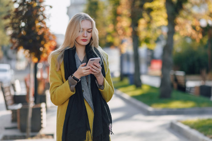 A woman in a yellow coat and black scarf looks at her phone, standing on a tree-lined sidewalk. A woman in a yellow coat and black scarf looks at her phone, standing on a tree-lined sidewalk.