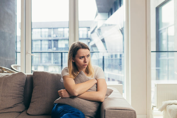 Concerned stepmom sitting on a couch, holding a pillow, pondering her role in her husband’s kids' lives. Concerned stepmom sitting on a couch, holding a pillow, pondering her role in her husband’s kids' lives.
