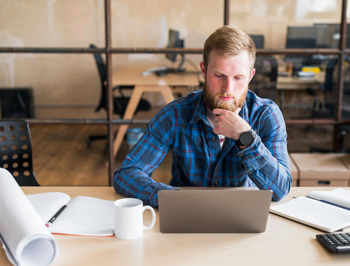 Man in a blue plaid shirt working at a laptop, surrounded by office items, appearing focused. Man in a blue plaid shirt working at a laptop, surrounded by office items, appearing focused.
