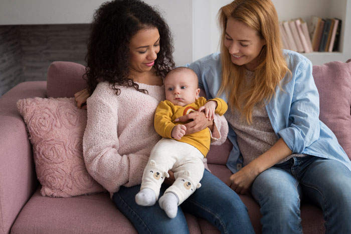 Two women smiling at a baby sitting on their lap, celebrating a friend's birthday together on a pink sofa. Two women smiling at a baby sitting on their lap, celebrating a friend's birthday together on a pink sofa.