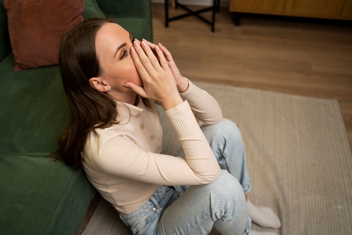 Woman sitting on floor, covering face, in a home setting with chaotic background and unclean surfaces. Woman sitting on floor, covering face, in a home setting with chaotic background and unclean surfaces.