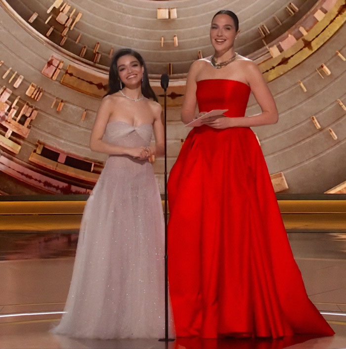 Two women elegantly dressed on stage at an event, one in a silver gown and the other in red. Two women elegantly dressed on stage at an event, one in a silver gown and the other in red.