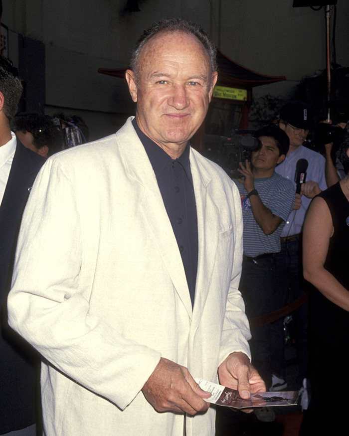Gene Hackman in a white jacket at an event, holding a paper, surrounded by people and cameras. Gene Hackman in a white jacket at an event, holding a paper, surrounded by people and cameras.