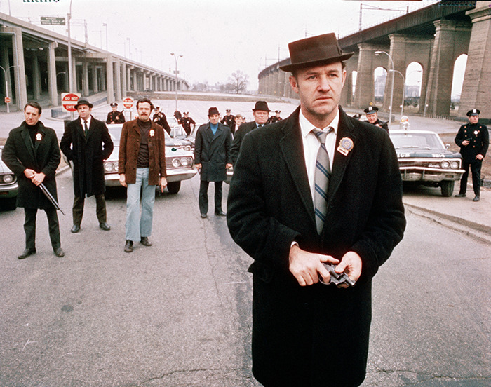 Gene Hackman in a classic film scene wearing a hat and coat, looking serious, with vintage police cars behind him. Gene Hackman in a classic film scene wearing a hat and coat, looking serious, with vintage police cars behind him.