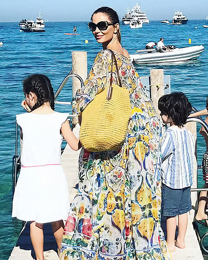 Georgina Chapman in a colorful dress and sunglasses, holding hands with two children on a pier, with boats in the background. Georgina Chapman in a colorful dress and sunglasses, holding hands with two children on a pier, with boats in the background.
