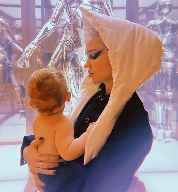 Child held by a person in a white hood, with reflective robotic figures in the background. Child held by a person in a white hood, with reflective robotic figures in the background.