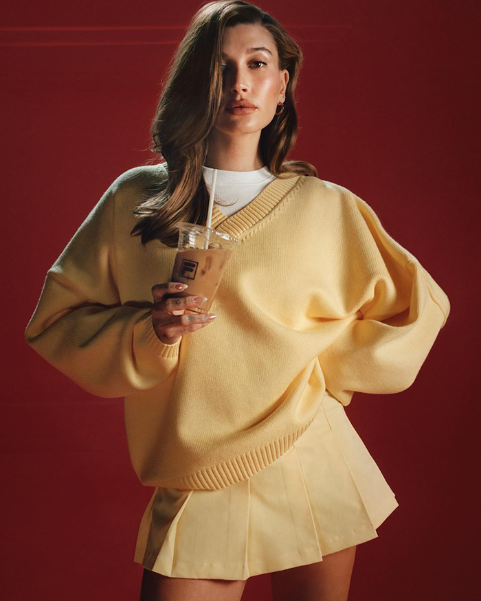 Hailey Bieber in yellow sweater holding iced coffee, posing against red backdrop. Hailey Bieber in yellow sweater holding iced coffee, posing against red backdrop.