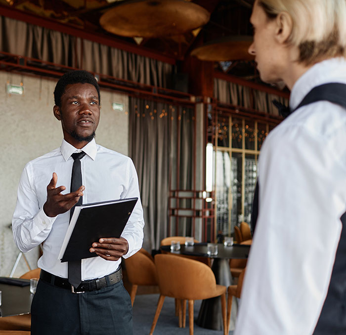 Two employees in uniform talking in a restaurant setting, holding a clipboard. Two employees in uniform talking in a restaurant setting, holding a clipboard.