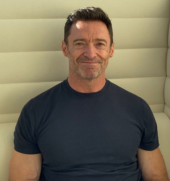 Hugh Jackman in a casual setting, seated and smiling, wearing a dark t-shirt. Hugh Jackman in a casual setting, seated and smiling, wearing a dark t-shirt.