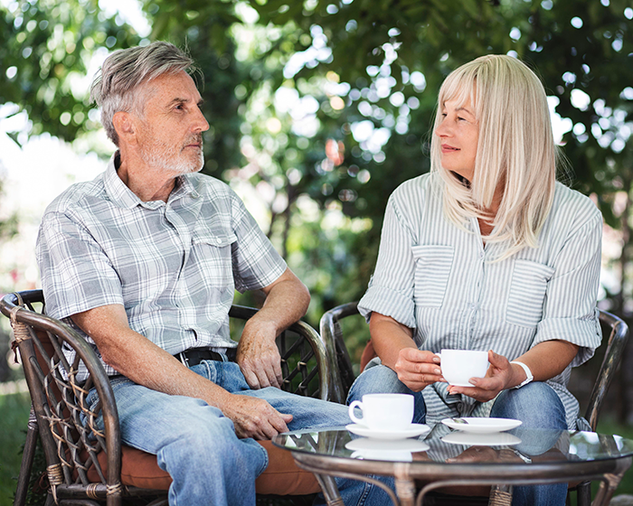 A husband and wife sitting outdoors, enjoying coffee together, surrounded by leafy trees. A husband and wife sitting outdoors, enjoying coffee together, surrounded by leafy trees.
