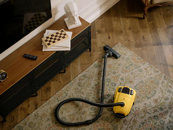 Yellow vacuum cleaner on a floral rug next to a TV stand with books and a chessboard; husband gift idea. Yellow vacuum cleaner on a floral rug next to a TV stand with books and a chessboard; husband gift idea.