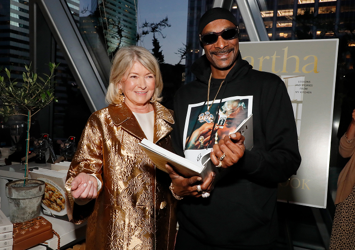 Snoop Dogg holding a book, smiling alongside a woman in a gold coat, highlighting his shift from rap royalty to business deals. Snoop Dogg holding a book, smiling alongside a woman in a gold coat, highlighting his shift from rap royalty to business deals.