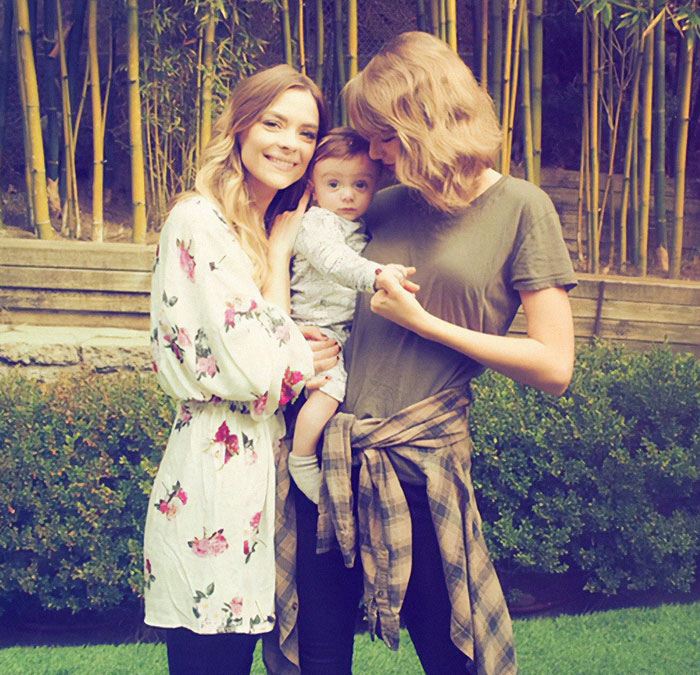 Two women outdoors holding a baby, one in floral, the other in a green shirt, representing Taylor Swift's godson situation. Two women outdoors holding a baby, one in floral, the other in a green shirt, representing Taylor Swift's godson situation.