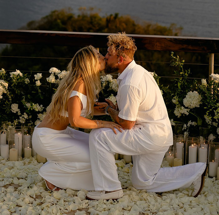 Couple embraces in a romantic proposal setting decorated with white flowers and candles, featuring Jake Paul. Couple embraces in a romantic proposal setting decorated with white flowers and candles, featuring Jake Paul.