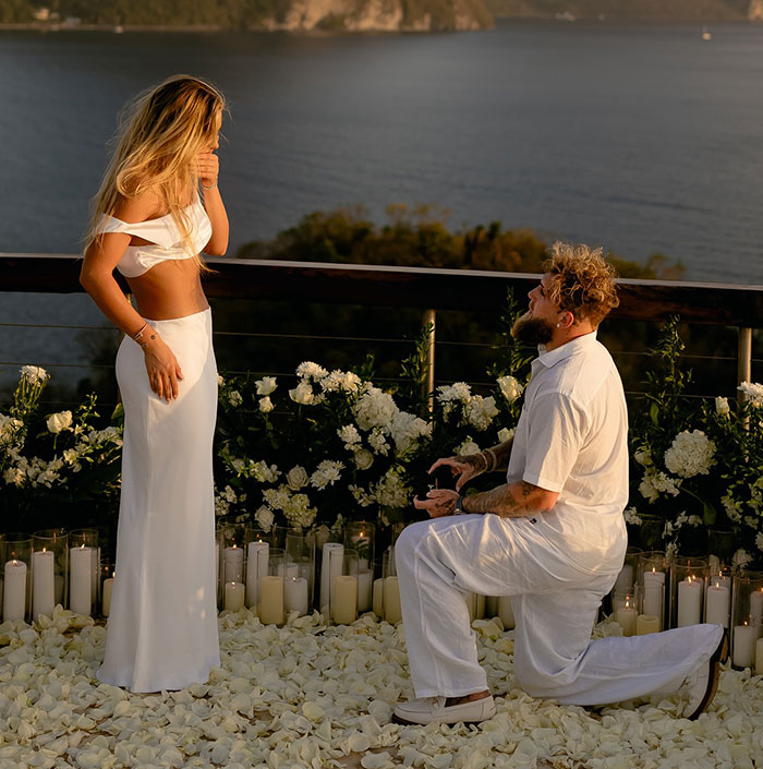 Man proposing to woman on a deck with flowers and candles, overlooking a scenic ocean view at sunset. Man proposing to woman on a deck with flowers and candles, overlooking a scenic ocean view at sunset.