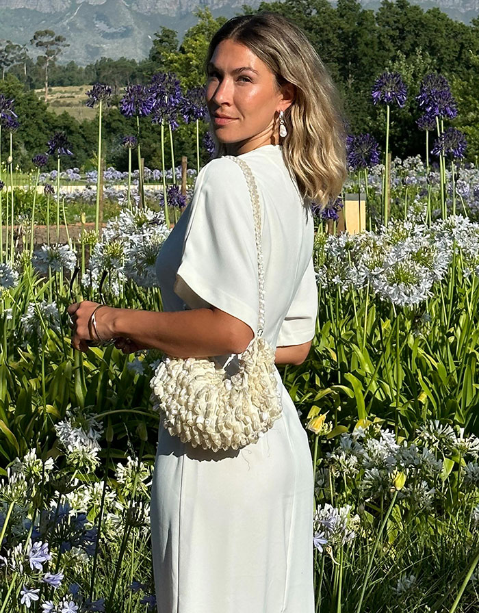 Woman in elegant white dress standing in a garden, holding a textured bag, with purple flowers in the background. Woman in elegant white dress standing in a garden, holding a textured bag, with purple flowers in the background.
