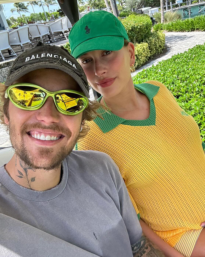 Hailey Bieber and Justin Bieber taking a selfie outdoors, one wearing a yellow shirt and green cap, the other in a gray shirt and sunglasses. Hailey Bieber and Justin Bieber taking a selfie outdoors, one wearing a yellow shirt and green cap, the other in a gray shirt and sunglasses.