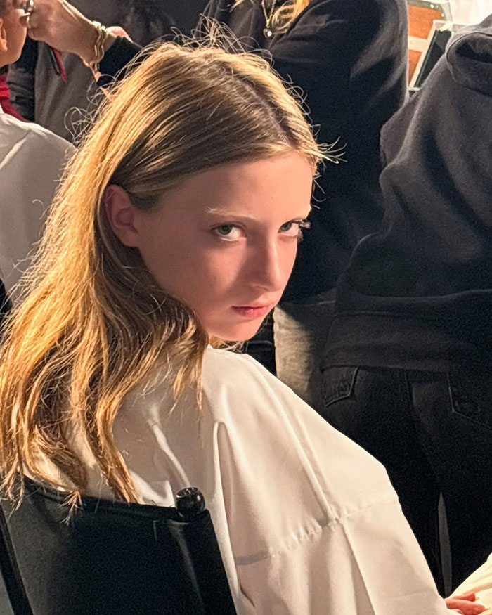 Young model at Paris Fashion Week, sitting in a chair, wearing a white outfit, hair styled, surrounded by people in the backstage. Young model at Paris Fashion Week, sitting in a chair, wearing a white outfit, hair styled, surrounded by people in the backstage.