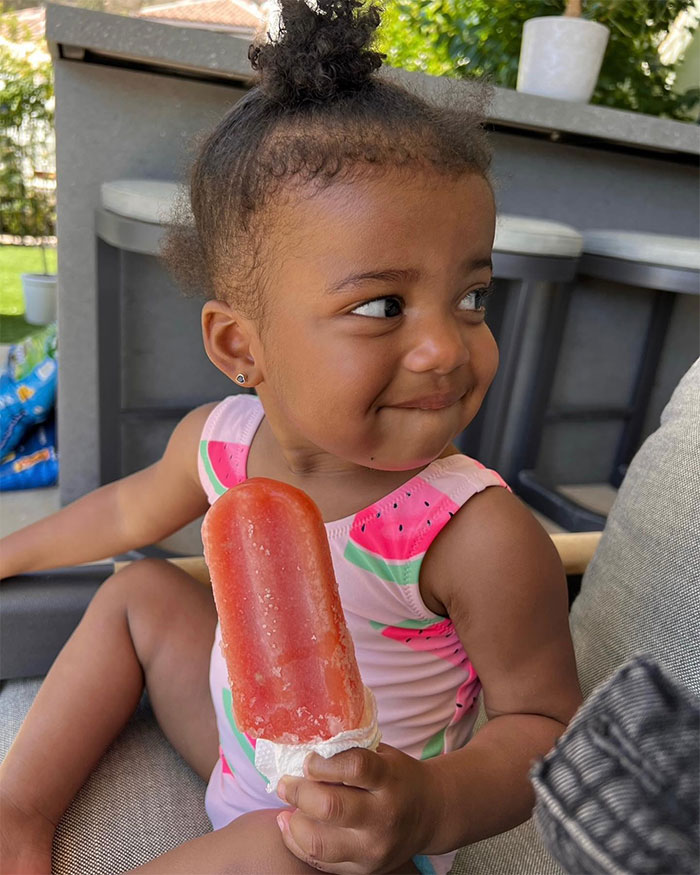 Toddler in a watermelon outfit enjoying a red popsicle, smiling outdoors. Toddler in a watermelon outfit enjoying a red popsicle, smiling outdoors.