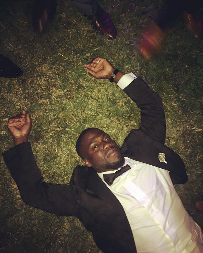Kevin Hart in a tuxedo lying on grass with arms outstretched, wearing a bow tie and watch, at an outdoor event. Kevin Hart in a tuxedo lying on grass with arms outstretched, wearing a bow tie and watch, at an outdoor event.