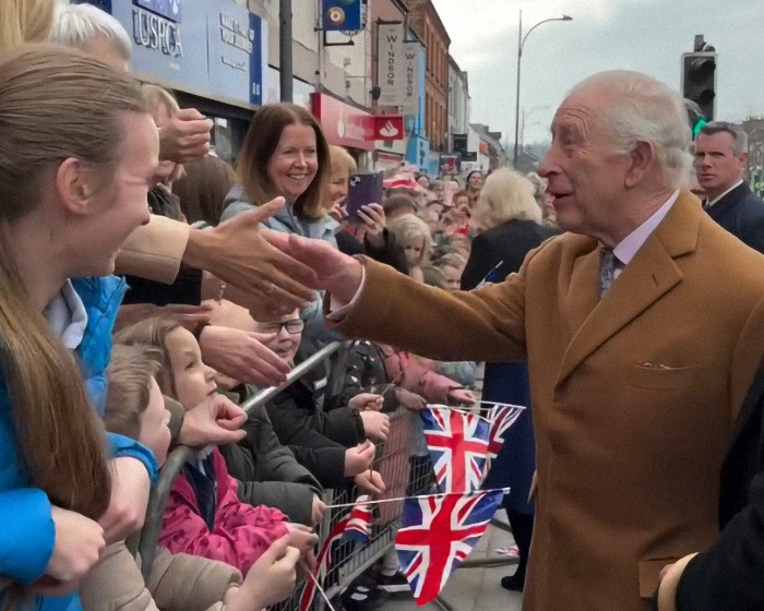 A royal figure greets fans; community shows support amid cancer battle, flags visible in the background. A royal figure greets fans; community shows support amid cancer battle, flags visible in the background.