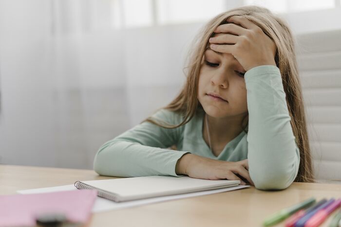 Young girl in a green sweater, bored while studying at a desk, represents a normal yet weird societal expectation.