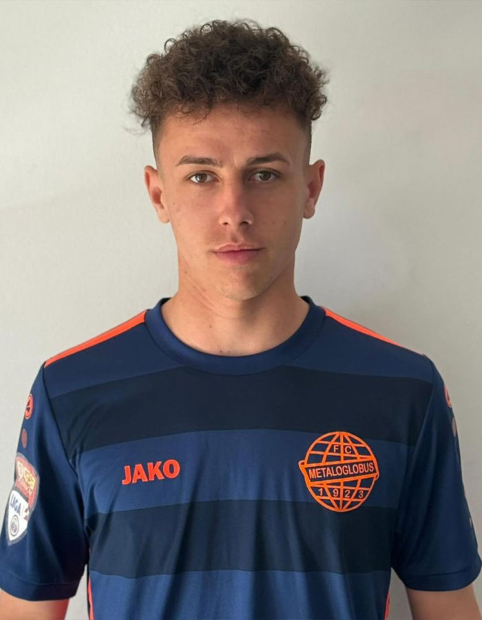 Young footballer in a blue and orange Metaloglobus jersey standing against a plain background. Young footballer in a blue and orange Metaloglobus jersey standing against a plain background.