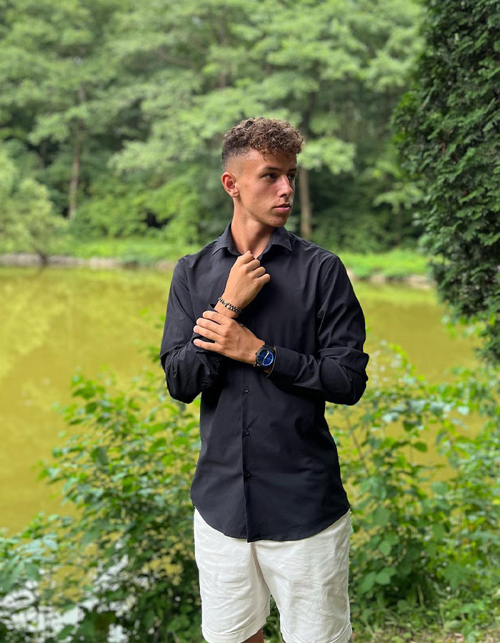 A 19-year-old footballer in a black shirt and white shorts stands beside a green, tree-lined pond. A 19-year-old footballer in a black shirt and white shorts stands beside a green, tree-lined pond.