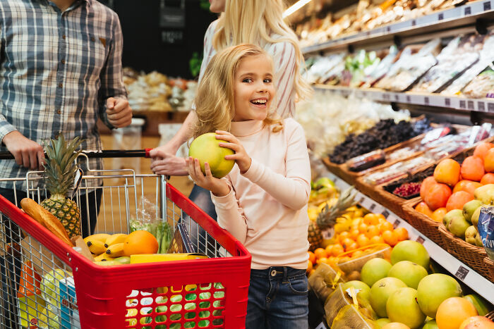 Child holding fruit, smiling at supermarket aisle with family. Child holding fruit, smiling at supermarket aisle with family.