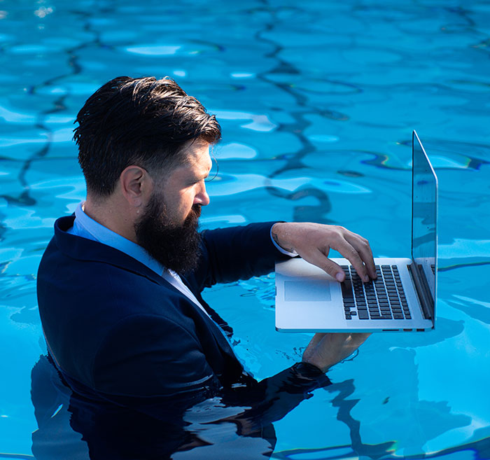 Man in a suit using a laptop while standing waist-deep in a pool, symbolizing work commitment even on vacation. Man in a suit using a laptop while standing waist-deep in a pool, symbolizing work commitment even on vacation.