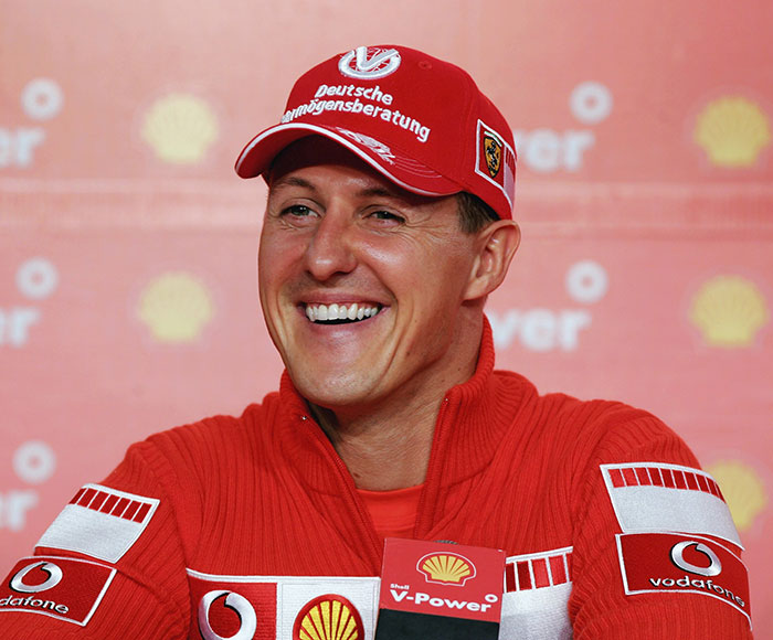 F1 legend in red cap and jacket, smiling at a press event, with Shell and Vodafone branding in the background. F1 legend in red cap and jacket, smiling at a press event, with Shell and Vodafone branding in the background.