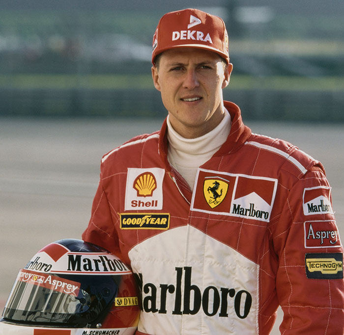Man in a red racing suit and cap holding a helmet, representing F1 legend Michael Schumacher. Man in a red racing suit and cap holding a helmet, representing F1 legend Michael Schumacher.