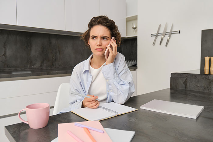 Woman in kitchen on phone, looking concerned, with notebook and coffee mug, considering wedding invitations expectations. Woman in kitchen on phone, looking concerned, with notebook and coffee mug, considering wedding invitations expectations.