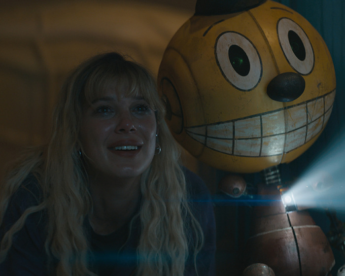 Blonde woman with an excited expression next to a large yellow robot, emitting light. Blonde woman with an excited expression next to a large yellow robot, emitting light.