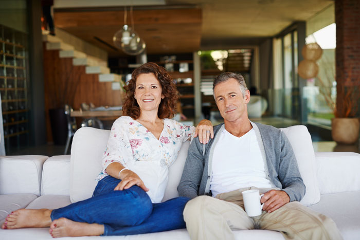 Couple sitting on a white couch in a modern living room, lady in floral blouse and jeans, man holding a coffee cup. Couple sitting on a white couch in a modern living room, lady in floral blouse and jeans, man holding a coffee cup.