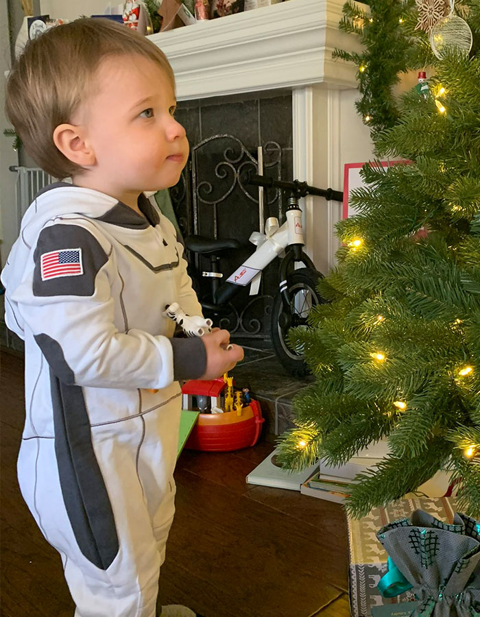 Toddler in a NASA-themed outfit, holding a toy, standing beside a lit Christmas tree indoors. Toddler in a NASA-themed outfit, holding a toy, standing beside a lit Christmas tree indoors.