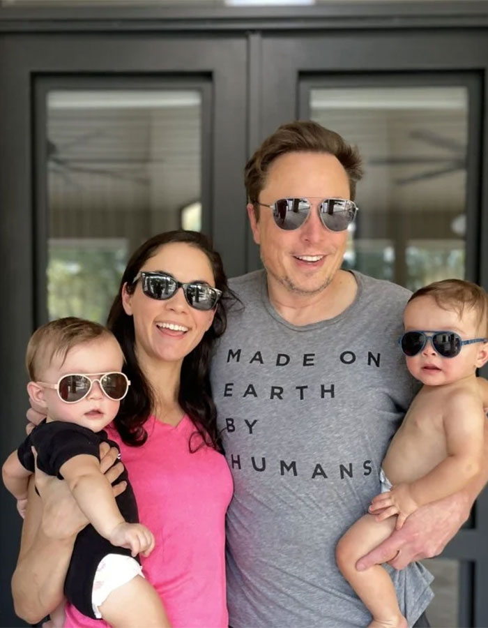 A man in a gray T-shirt and sunglasses stands with a woman in pink, holding two babies in sunglasses, smiling outdoors. A man in a gray T-shirt and sunglasses stands with a woman in pink, holding two babies in sunglasses, smiling outdoors.