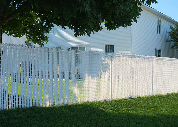 White fence casting shadows on grass, separating houses in a neighborhood with green trees and blue skies. White fence casting shadows on grass, separating houses in a neighborhood with green trees and blue skies.