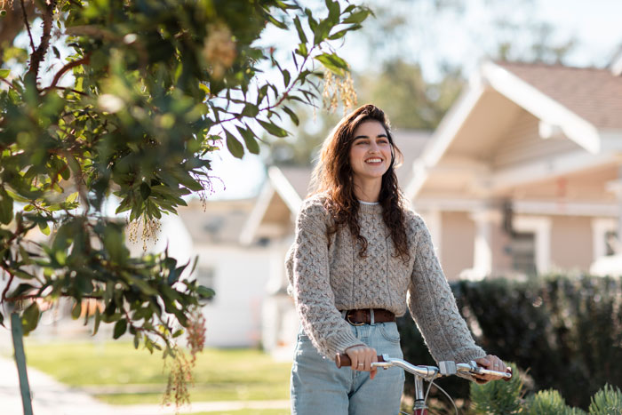 Woman smiling while biking in a neighborhood setting, wearing a knitted sweater and jeans, with greenery in the background. Woman smiling while biking in a neighborhood setting, wearing a knitted sweater and jeans, with greenery in the background.