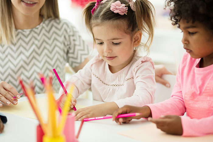Young kids coloring with pencils at a daycare table, guided by a caregiver. Young kids coloring with pencils at a daycare table, guided by a caregiver.