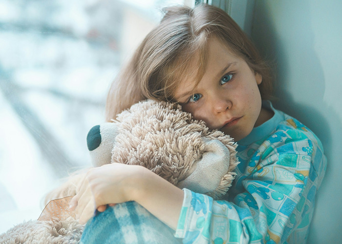 Child in pajamas hugging a teddy bear by a window, reflecting childhood experiences.