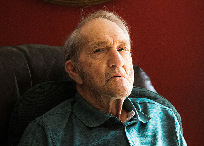 Elderly man sitting on a chair, wearing a green shirt, reflecting on childhood experiences.