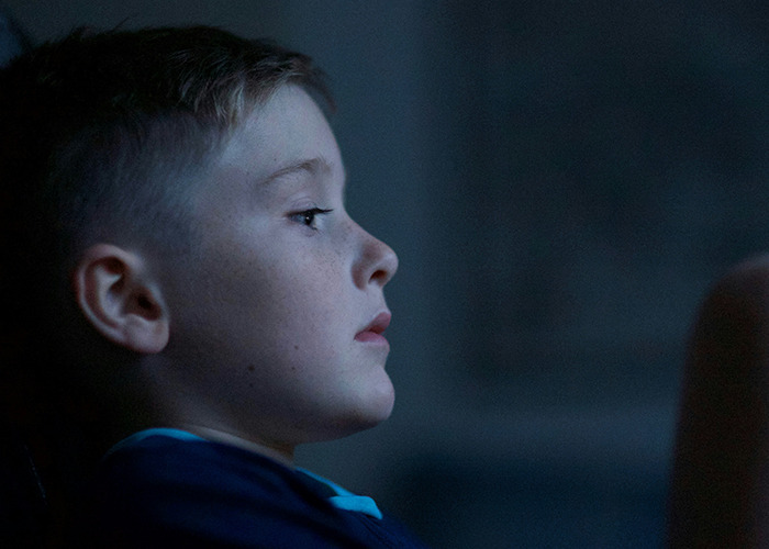 Young boy sitting in dim light, appearing thoughtful, illustrating childhood experiences that might be traumatic.