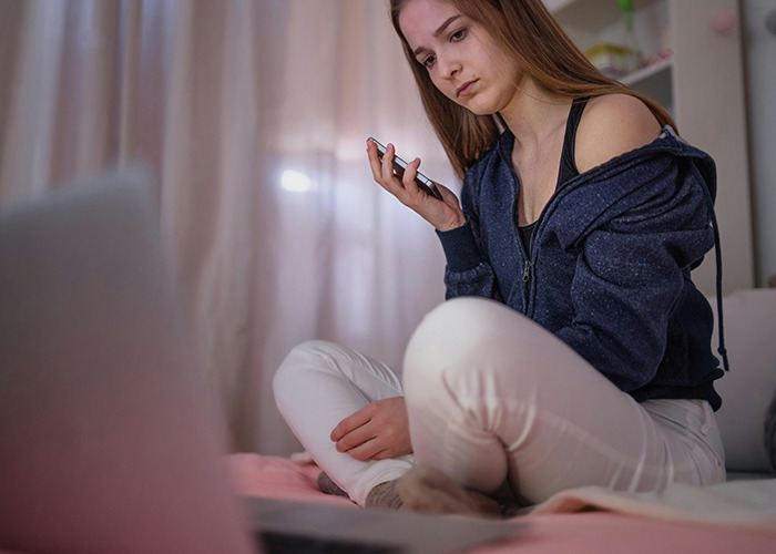 A young woman looking at a laptop, holding a phone, possibly reflecting on past childhood experiences.