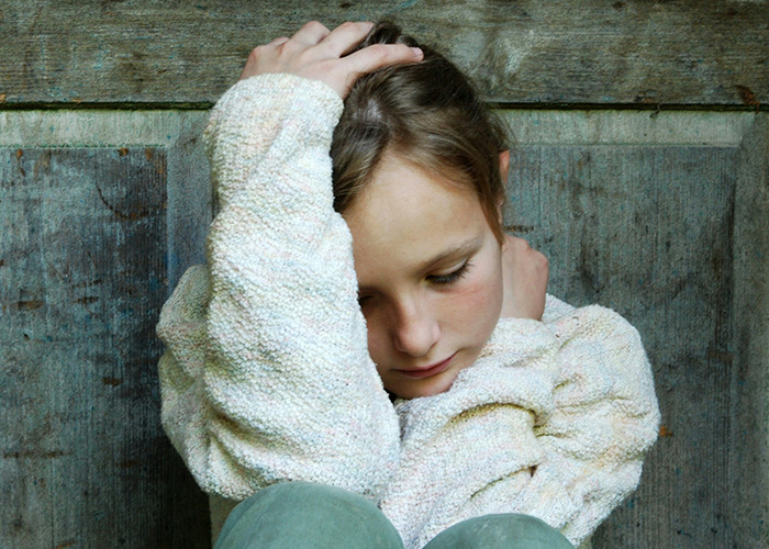 Child sitting against wooden wall, looking down, evokes complex childhood experiences.