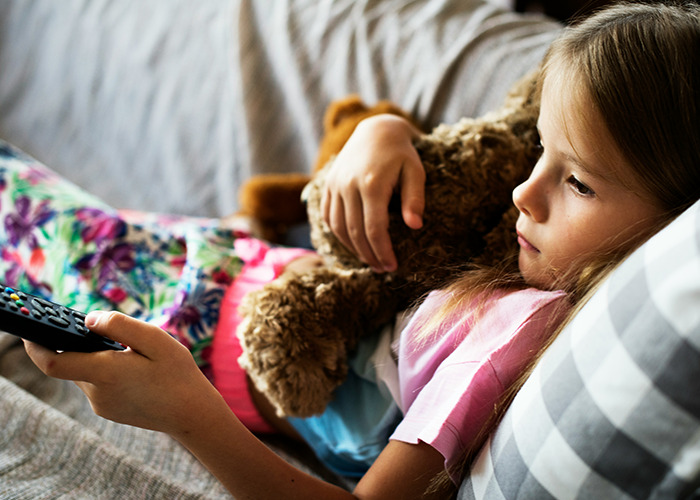 Young girl holding a bear and remote, lying on a couch, reflecting on childhood and traumatic experiences.