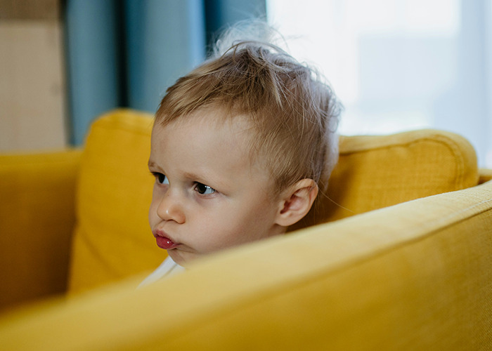 Child peering over a yellow couch, expressing curiosity, evoking childhood experiences and memories.