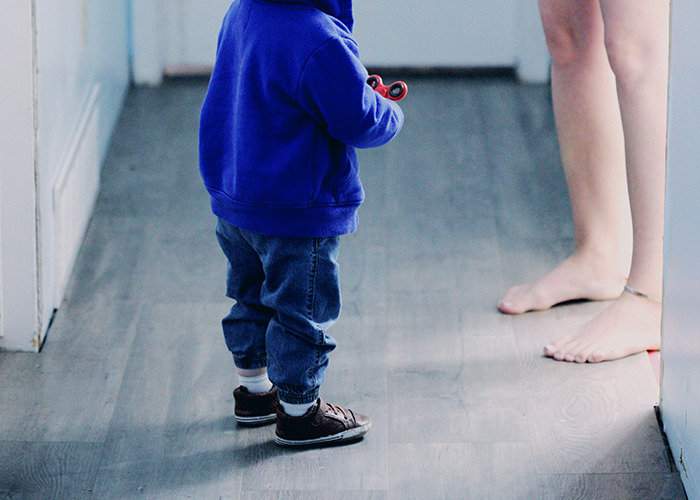 Toddler in blue jacket holding a toy, facing an adult's bare feet in a hallway, highlighting childhood experiences.