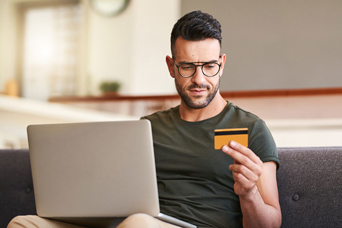 Man in glasses using laptop and holding credit card, reflecting on financial habits embraced by older generations.
